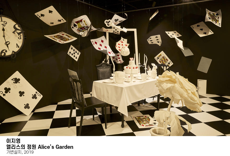 흰색천을 깐 테이블 위에 촛대, 컵, 케이크가 놓여져 있고 그 주변 천장에는 마치 트럼프카드가 휘날리는 듯 생동감 있게 매달려 있다. 바닥은 흰검의 격자무늬로 되어 있는 모습이다. 이미지 하단에는 이지영 앨리스의 정원 Alice's Garden, 가변설치, 2019 라고 쓰여져 있다