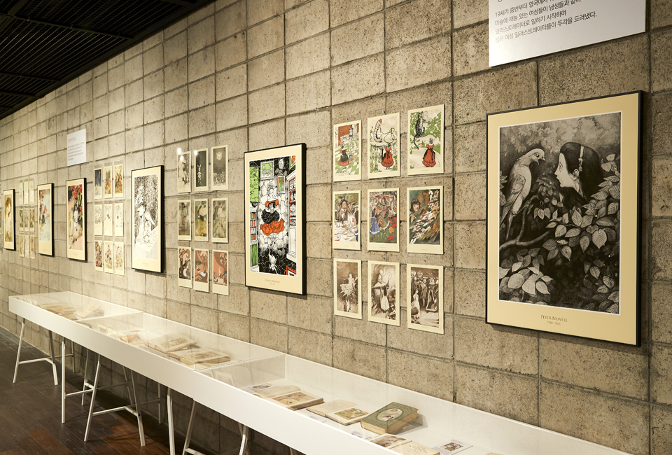벽면에 다양한 색감의 삽화들이 붙여져 있고, 그 아래 흰색 테이블 안에 책들이 펼쳐져 있는 상태로 전시되어 있는 모습을 찍은 사진이다.